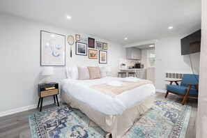 A modern bedroom with a large bed, a bedside table, a chair, and a kitchen area visible through an open door.