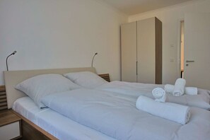 Uma cama de casal com lençóis brancos, uma mesa de cabeceira de madeira e um guarda-roupa com espelho.