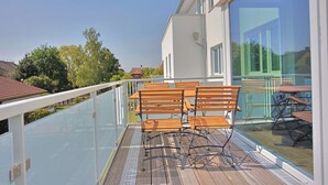 A balcony with wooden benches and a glass railing, overlooking a residential area with trees and houses. - Villa Baltic Star Ferienwohnung 10 (Börgerende-Rethwisch)
