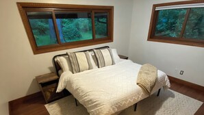 Een slaapkamer met een bed, een nachtkastje, een raam met uitzicht op bomen en een plafondlamp.