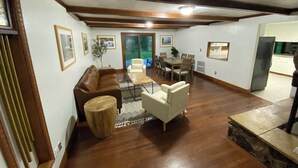 Een ruime woonkamer met een bruine leren bank, een witte fauteuil en een houten salontafel. Er is een eettafel met stoelen en een keuken te zien op de achtergrond.