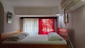 Een enkel bed met een houten hoofdbord, witte beddengoed en een rood gordijn. Er is een luchtverkoeler aan het plafond en een ingelijst beeld op de muur.