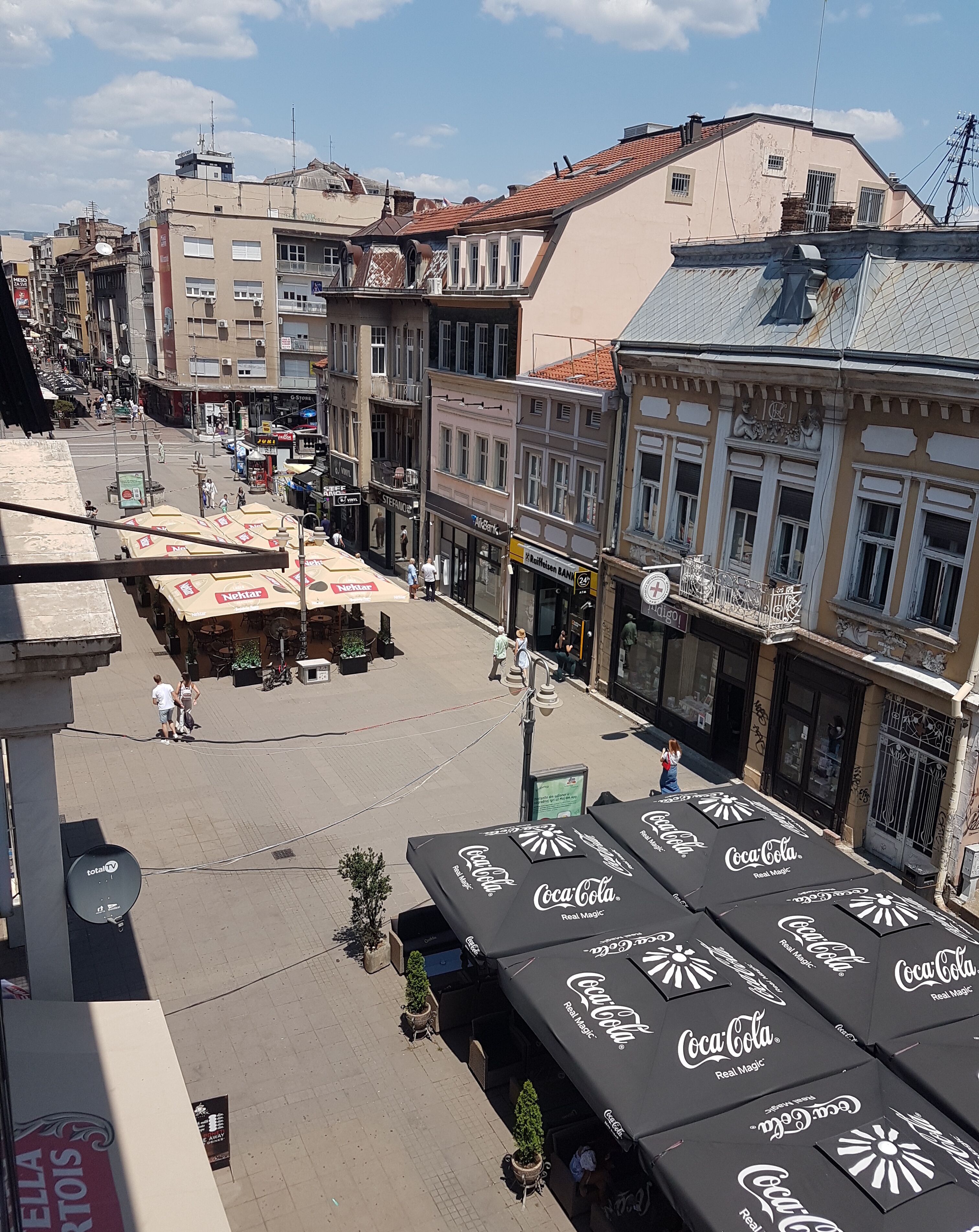 Vista de una calle con zonas de estar al aire libre cubiertas por sombrillas con la marca Coca-Cola, diversas tiendas y una combinación de edificios residenciales y comerciales.
