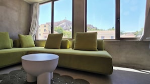 A modern living room with a green sofa, a white coffee table, and large windows offering a view of buildings and a flag. - 99 Adrianou Luxury Urban Stay (Athens)