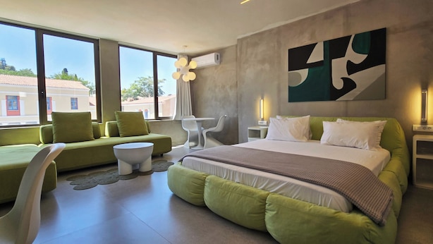 A modern hotel room with a large bed, green sofas, a white chair, and a view of buildings through large windows. - 99 Adrianou Luxury Urban Stay (Athens)