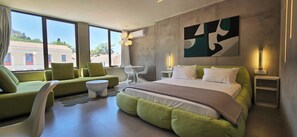 A modern hotel room with a large bed, green sofas, a white chair, and a view of buildings through large windows. - 99 Adrianou Luxury Urban Stay (Athens)