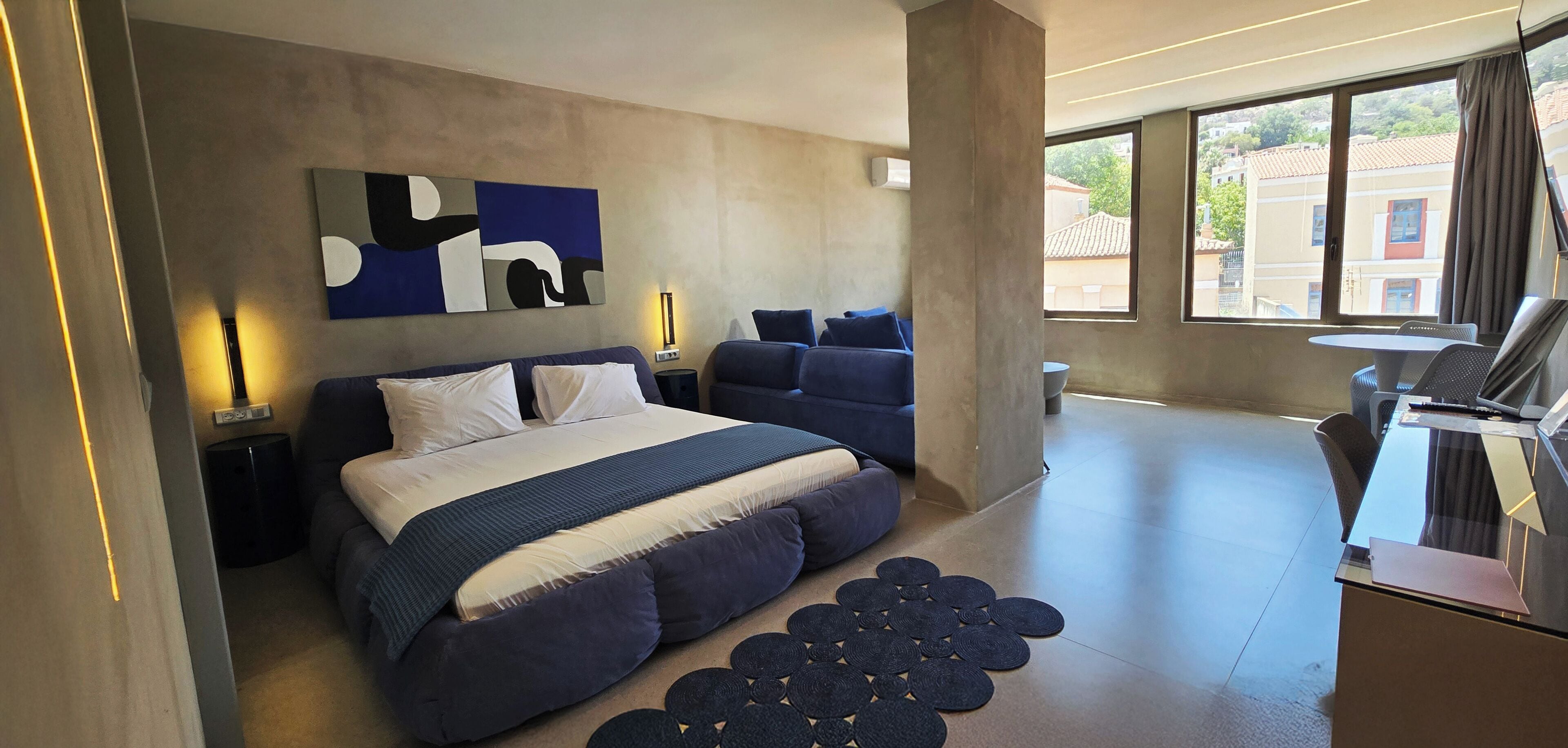 A modern bedroom with a large bed, a sofa, a desk, and a view of buildings outside.