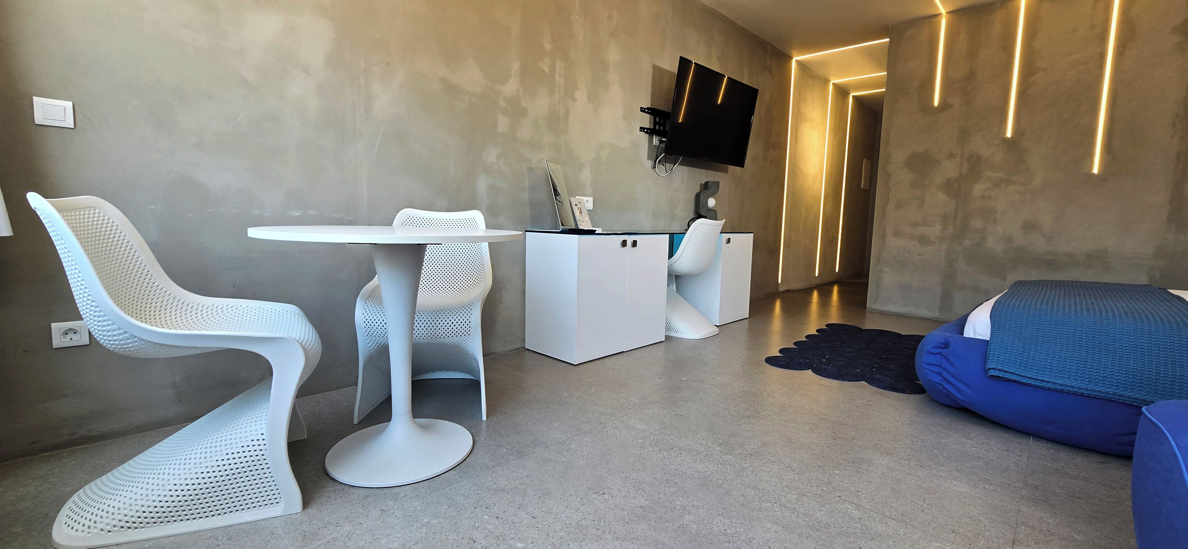 A modern interior with a white chair, a small round table, a desk with a chair, a TV mounted on the wall, and a blue sofa.