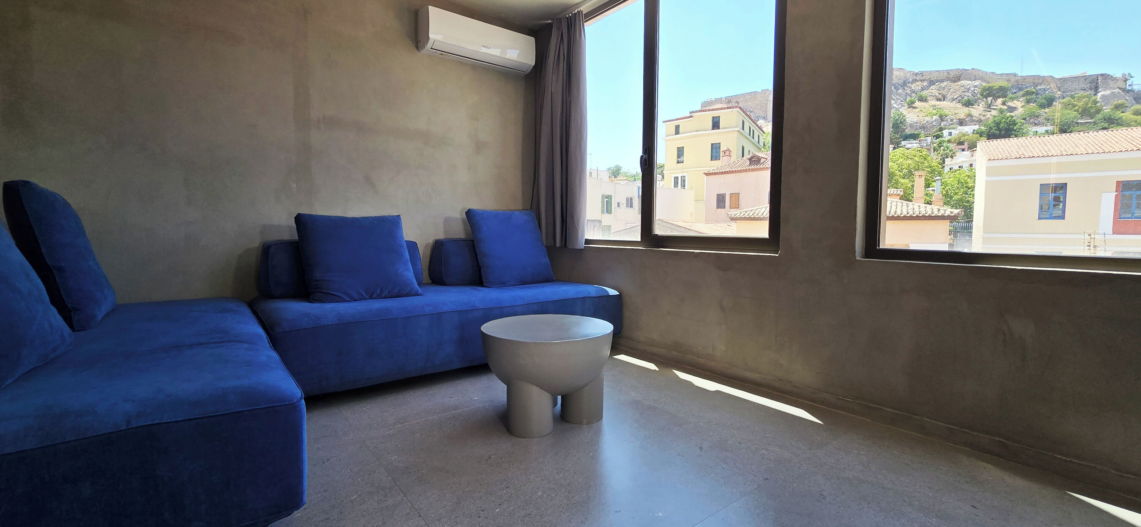 Una stanza moderna con un divano blu, un tavolino rotondo e un'ampia finestra con vista su edifici e colline.
