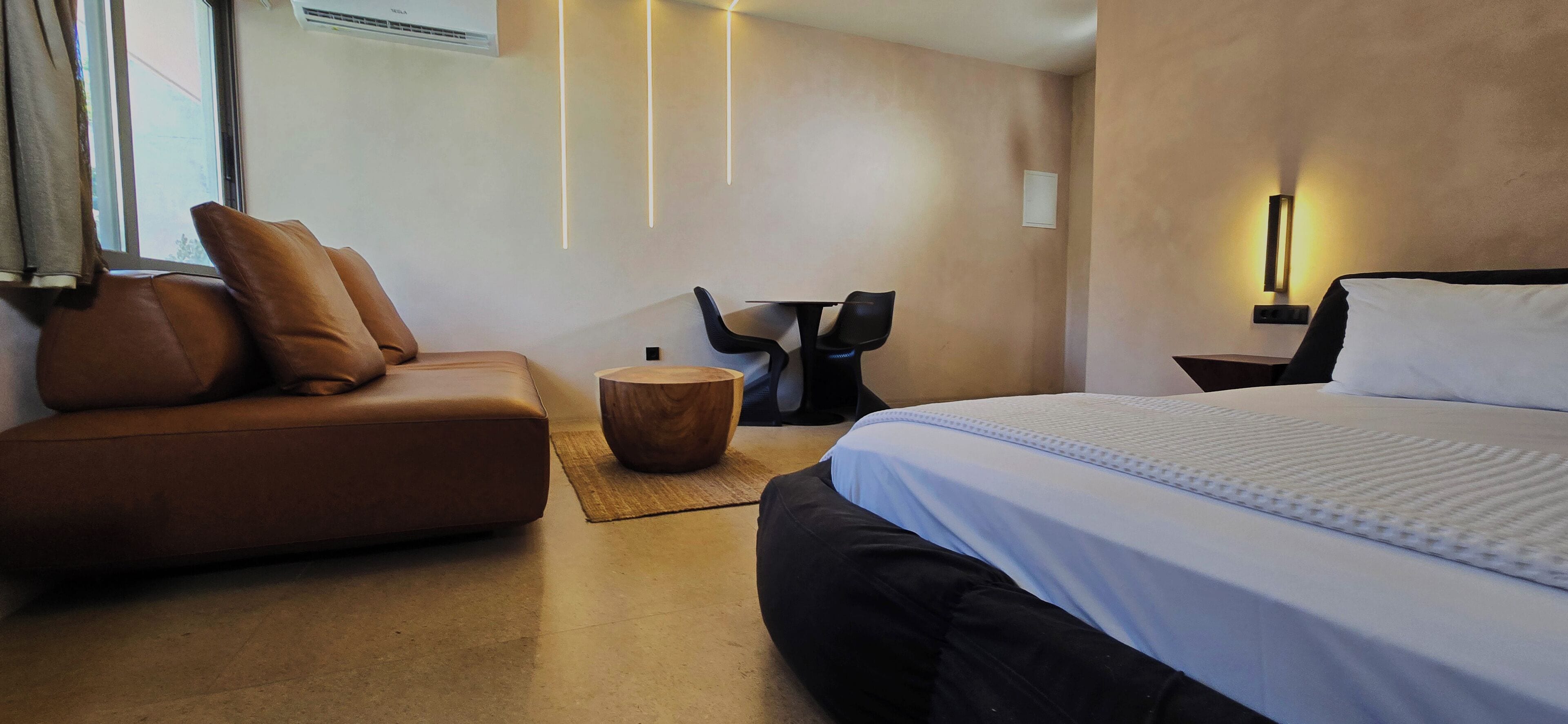 Una habitación de hotel moderna con una cama grande, un sofá de cuero marrón, una mesita redonda y un escritorio adosado a la pared.