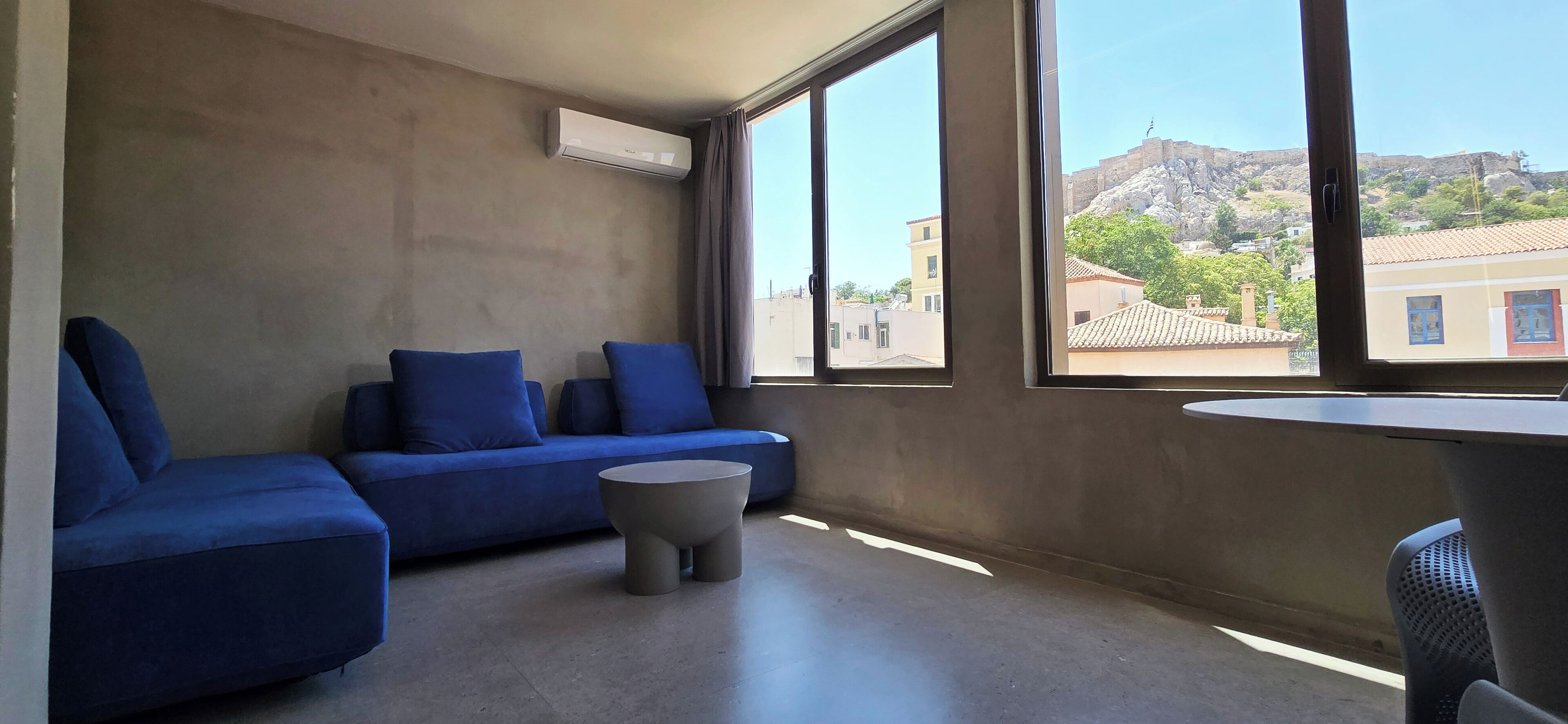 A modern room with a blue sofa, a small round table, and a large window offering a view of buildings and a hill.