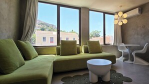 A modern living room with a green sofa, a white coffee table, and a view of buildings through large windows. - 99 Adrianou Luxury Urban Stay (Athens)
