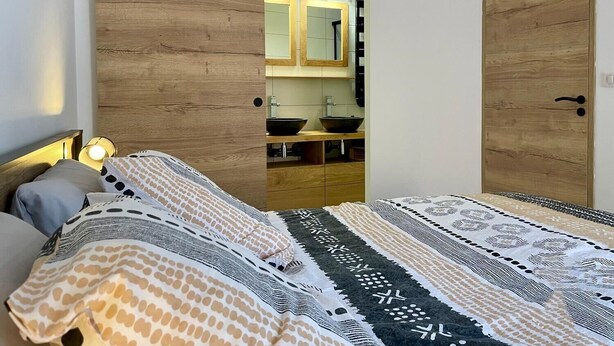 Une chambre à coucher avec un lit, une armoire en bois, une salle de bain avec un lavabo et un miroir, et une porte.