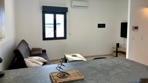 Un salon moderne avec une table grise, une planche à découper en bois dotée d’une spirale métallique, un canapé noir avec des coussins, une fenêtre donnant sur un paysage enneigé et un climatiseur fixé au mur.
