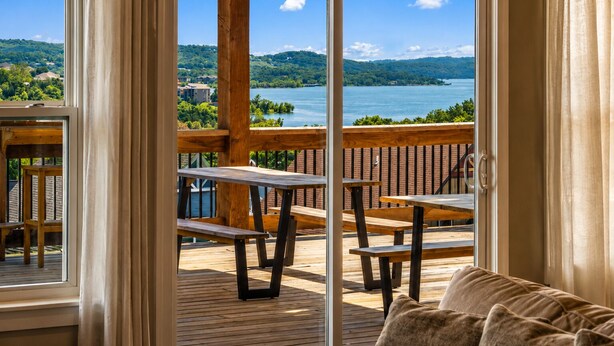 A deck with wooden benches and tables, a glass door leading to a view of a lake and hills.