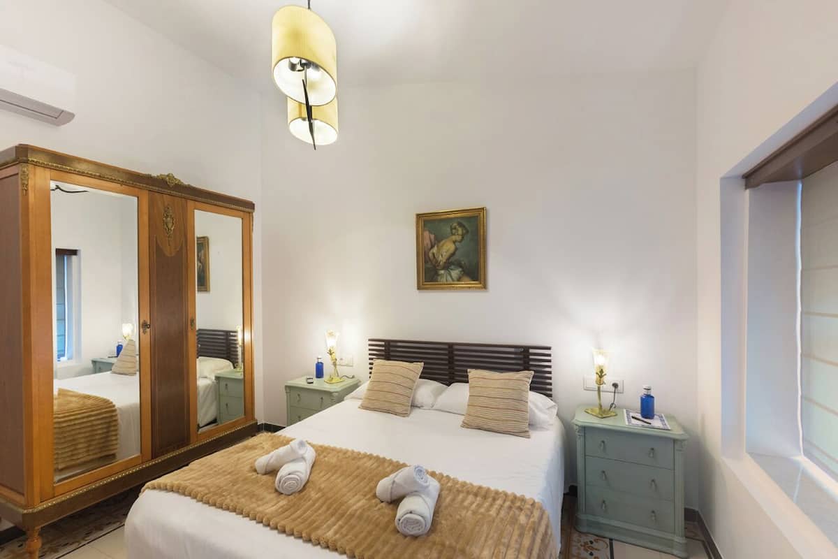 Een slaapkamer met een grote spiegel, een bed met witte lakens, twee nachtkastjes met lampen en een raam met zonwering.