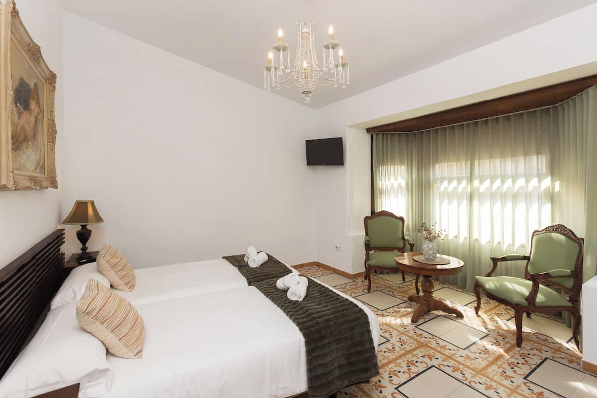 Een hotelkamer met een groot bed, een kroonluchter, een televisie, een eettafel met stoelen en een raam met gordijnen.