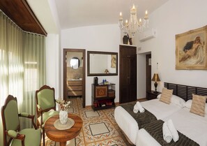 Een hotelkamer met een bed, een ronde houten tafel met een vaas, twee stoelen, een dressoir, een spiegel en een kroonluchter.