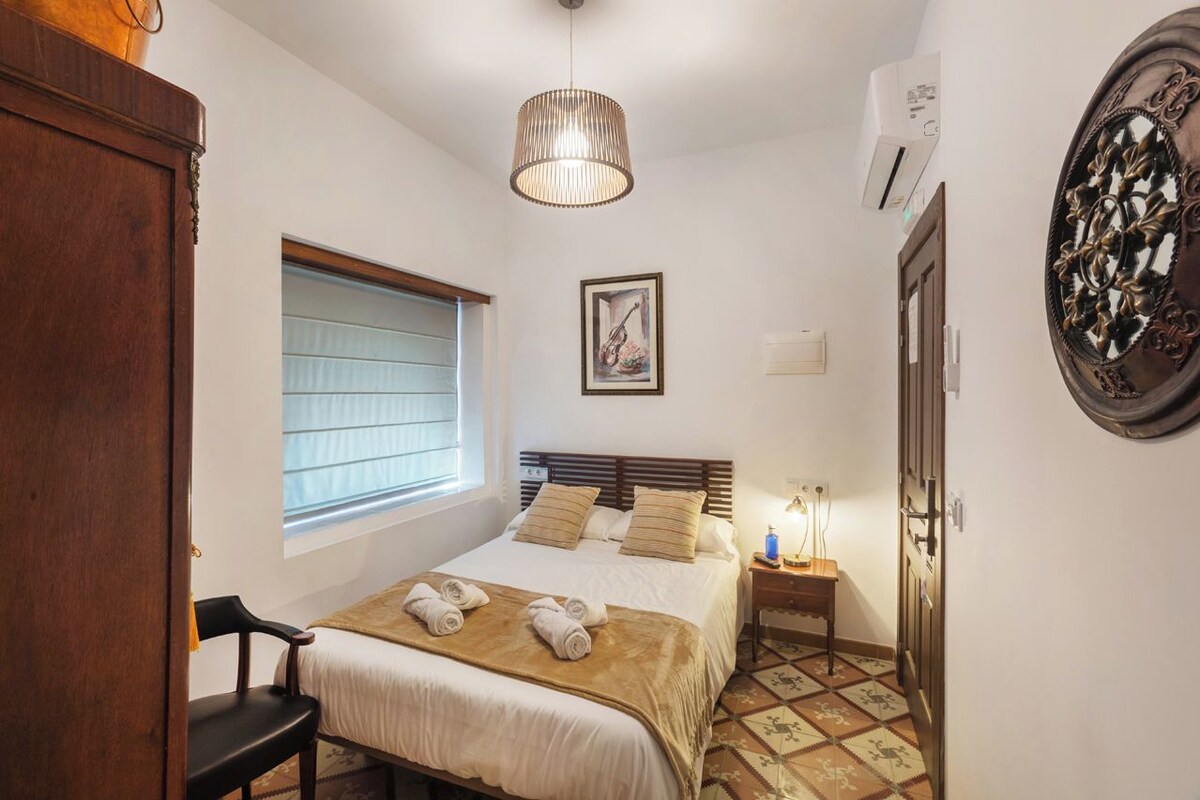 Un dormitorio con cama, una silla, una mesita de noche con lámpara, una ventana con persianas, un espejo montado en la pared y un suelo estampado.