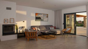 Un salon moderne avec un canapé, une table basse et deux fauteuils. On y trouve une cheminée, un tableau accroché au mur et une vue à travers une porte vitrée.