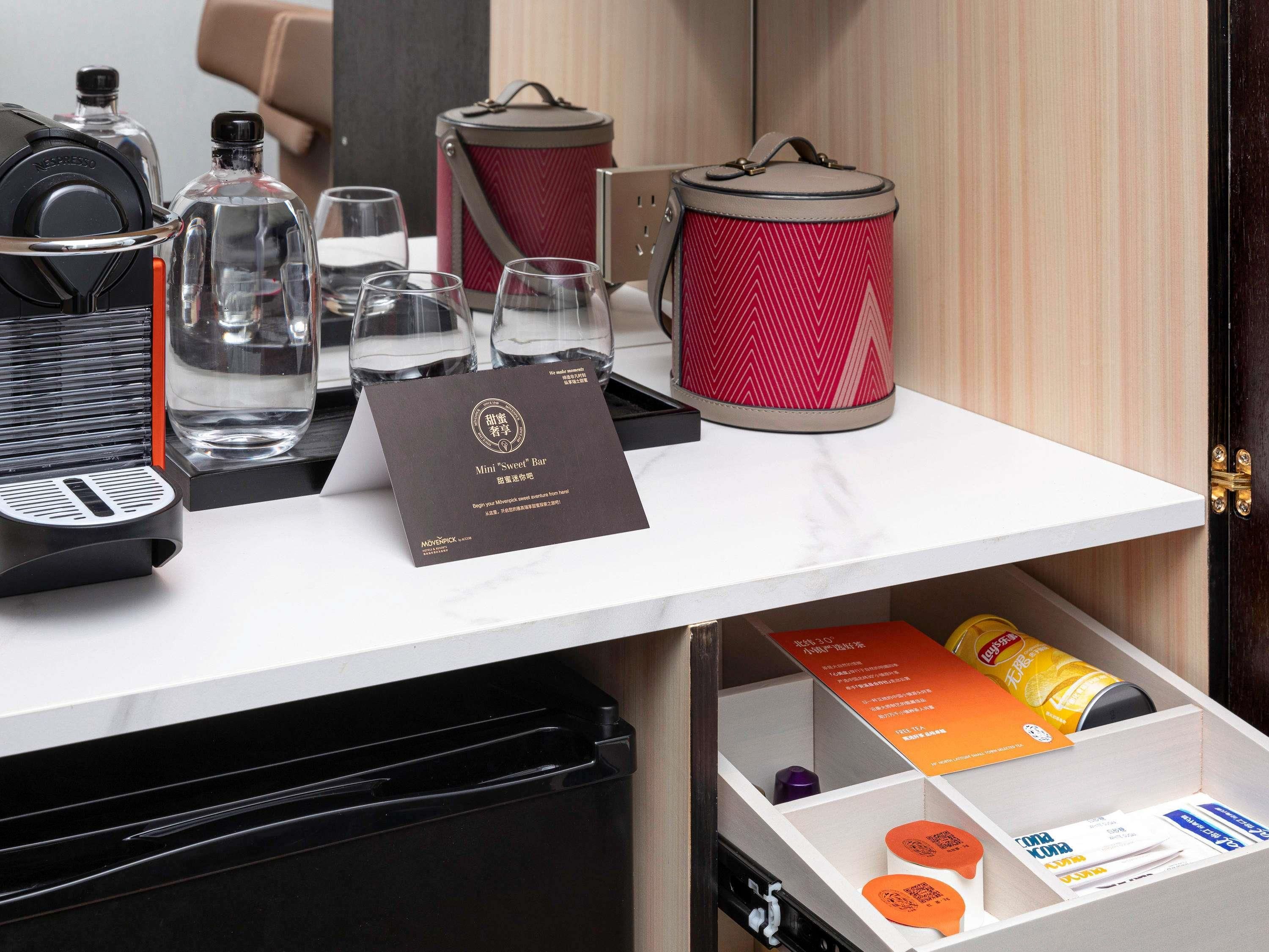 A minibar setup with a coffee machine, glasses, a red patterned container, and a card for a Mini "Sweet" Bar.