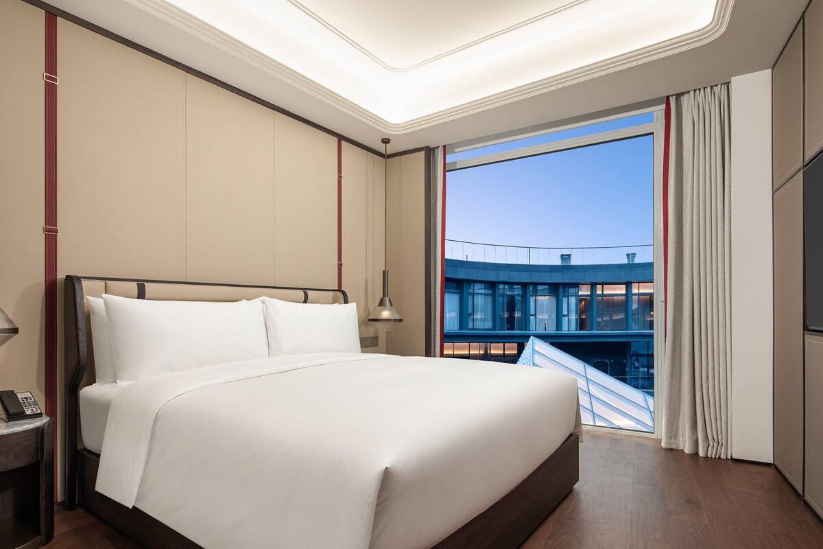 Hotelkamer met een groot bed, nachtkastje met telefoon, raam met gordijnen en uitzicht op een gebouw.