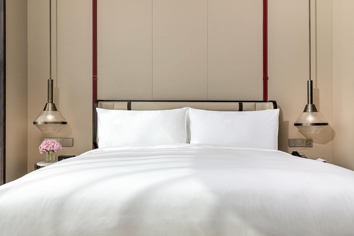 A neatly made bed with white linens, a headboard with a black frame, and two pendant lights hanging from the ceiling.