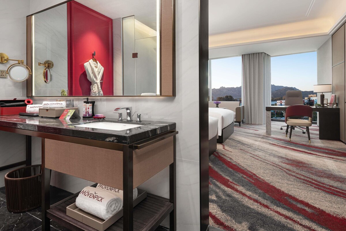 A hotel room with a large mirror, a vanity with a sink, a bed, a desk with a chair, and a view of mountains.