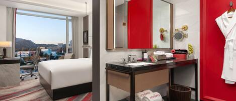 A hotel room with a bed, a desk, a bathroom with a mirror, and a view of the city.