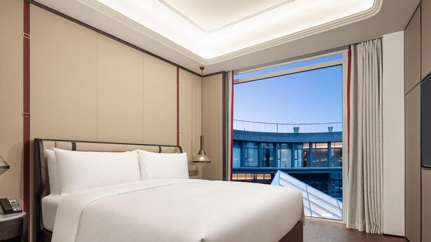 Ein modernes Hotelzimmer mit einem großen Bett, einem Nachttisch mit Telefon und Blick auf ein Gebäude durch ein Fenster.