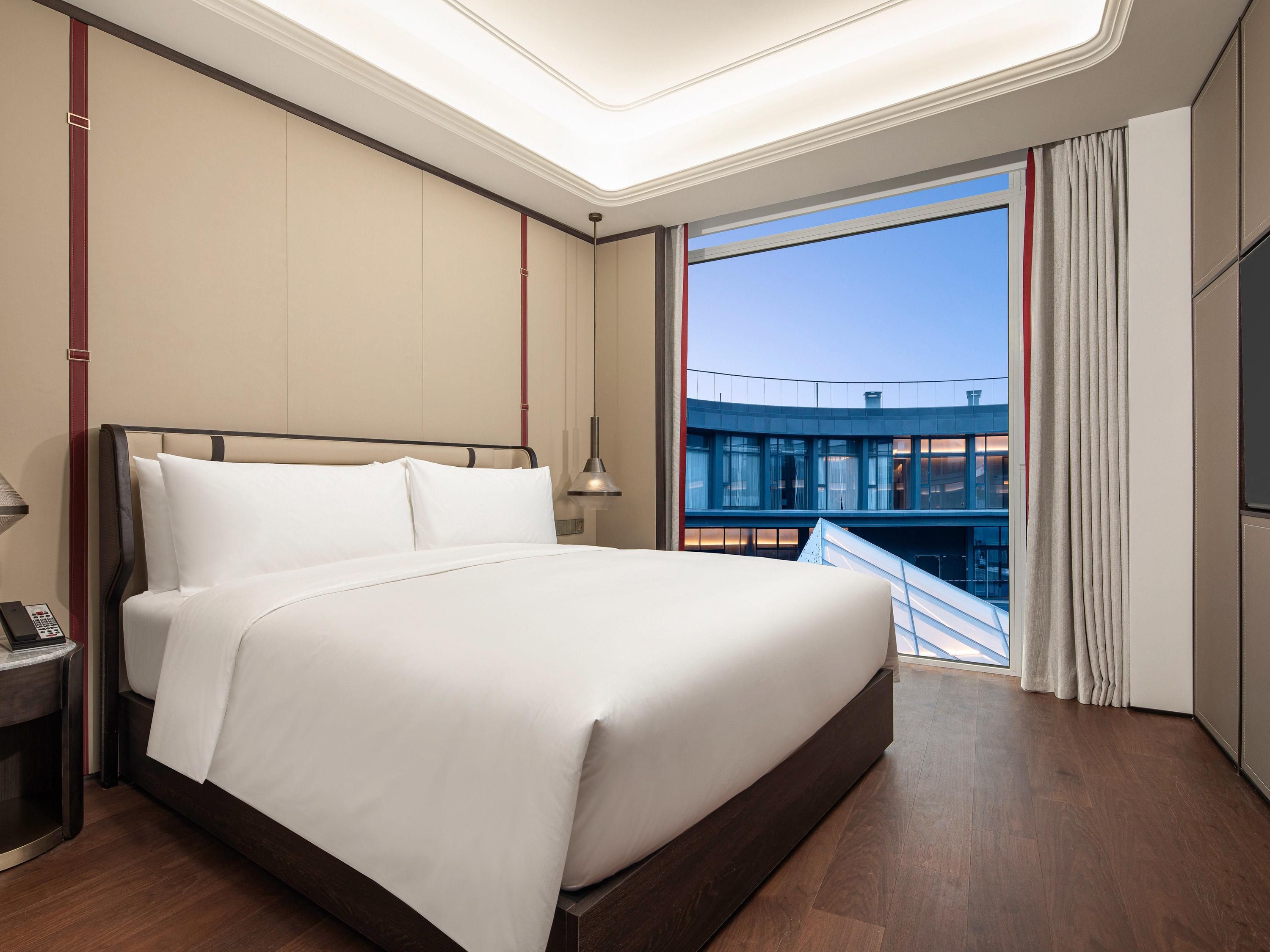 A hotel room with a large bed, a bedside table with a phone, a window with curtains, and a view of a building outside.
