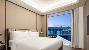Ein Hotelzimmer mit einem großen Bett, einem Nachttisch mit Telefon, einem Fenster mit Vorhängen und Blick auf ein Gebäude.