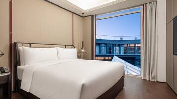 A hotel room with a large bed, a bedside table with a phone, a window with curtains, and a view of a building outside.