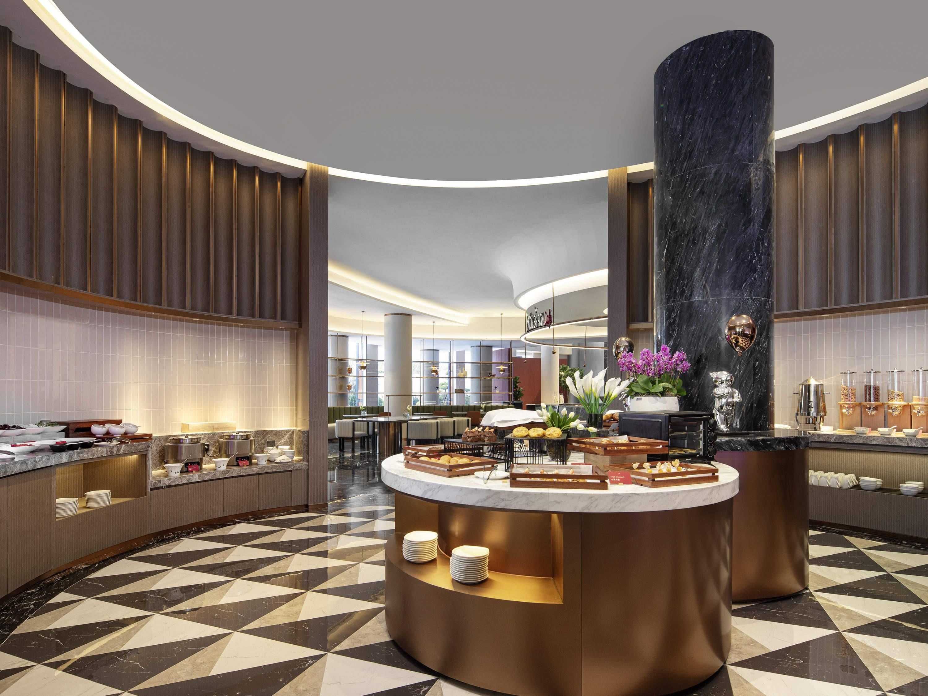A modern hotel lobby with a central round counter displaying breakfast items, surrounded by seating and a large pillar.