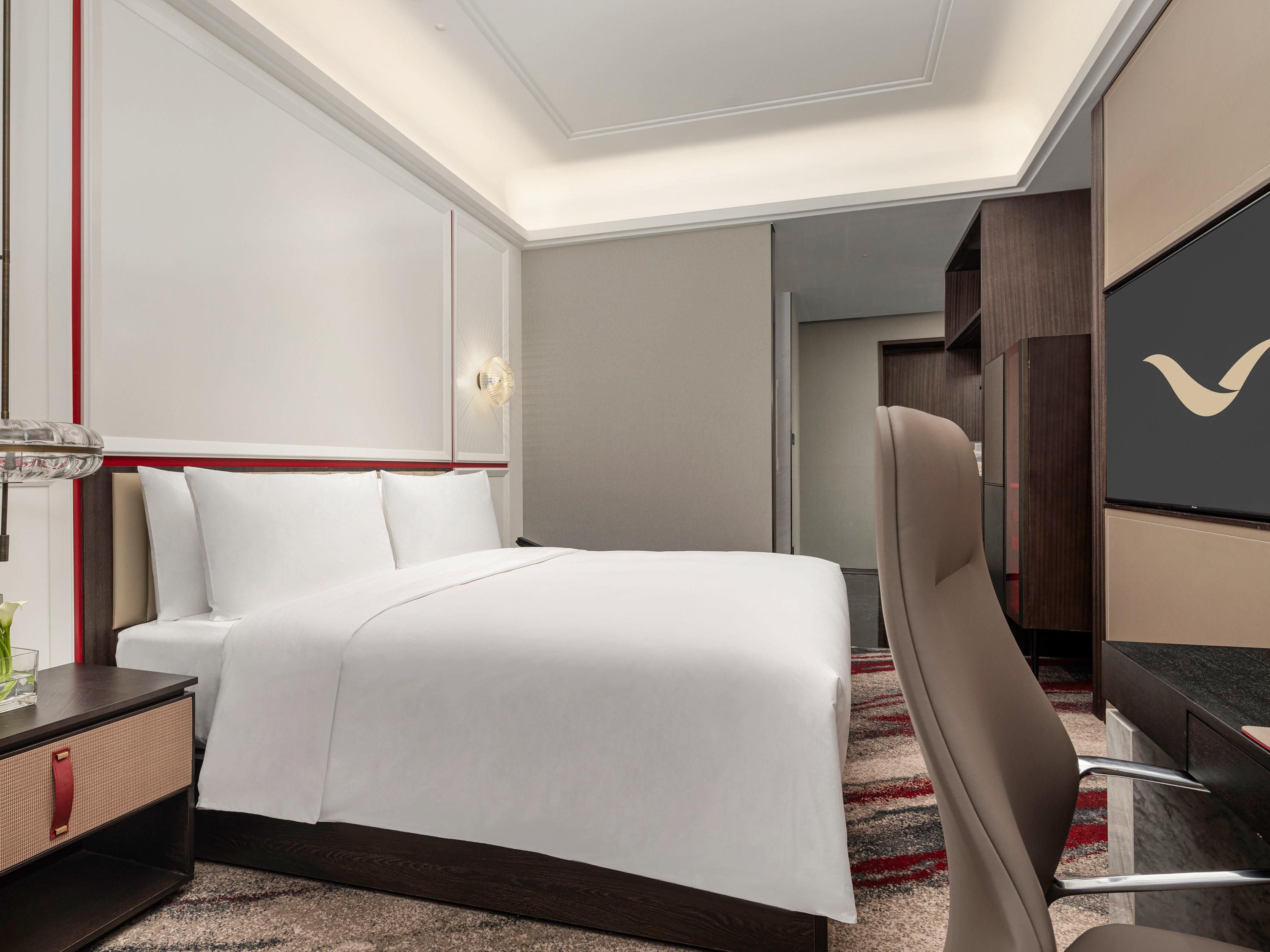 A hotel room with a neatly made bed, a desk with a chair, a TV, and a door leading to another room.