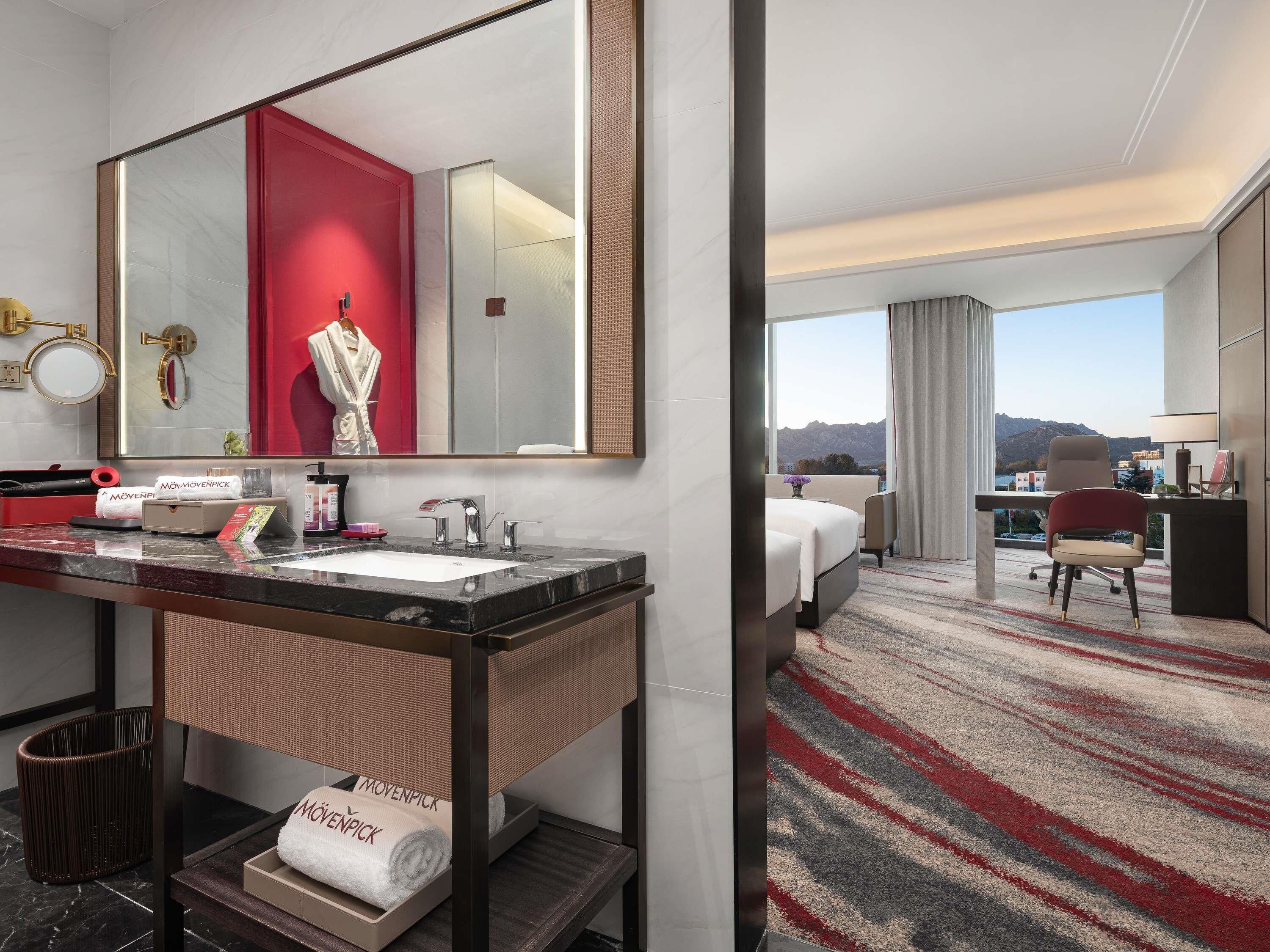 Hotelkamer met een grote spiegel, een wastafelmeubel met een wastafel, een bed, een bureau met een stoel en uitzicht op bergen.