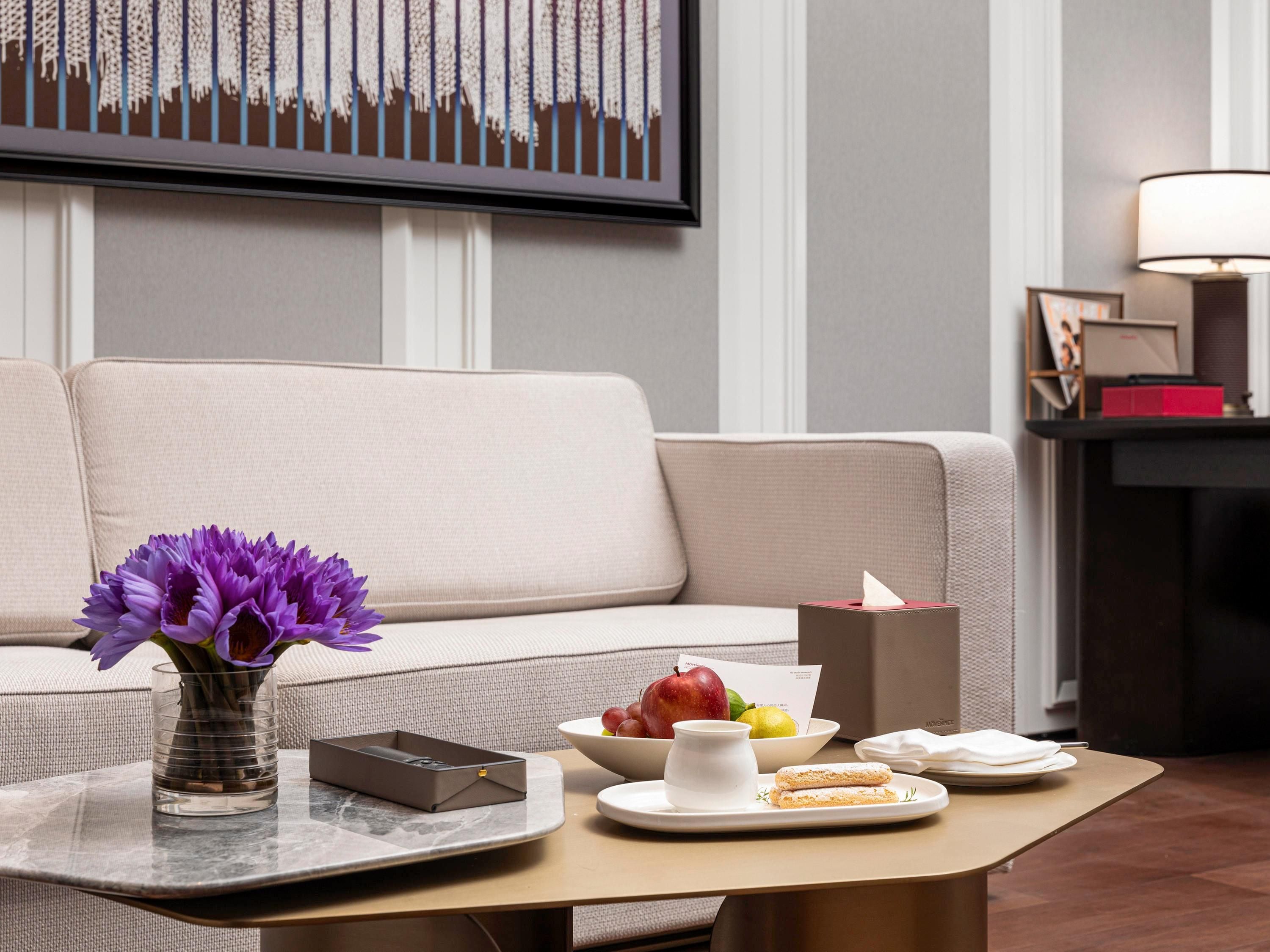 A modern living room with a sofa, a coffee table with a vase of flowers, a bowl of fruit, and a tray with a teapot and cups.