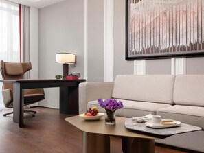 Een moderne hotelkamer met een bank, een bureau met een lamp, een salontafel met een schaal fruit en bloemen, en een kunstwerk aan de muur.