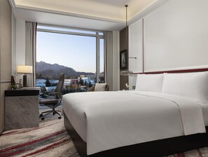 Camera d'albergo con un letto grande, una scrivania con una lampada, una sedia e vista sulle montagne e sugli edifici.