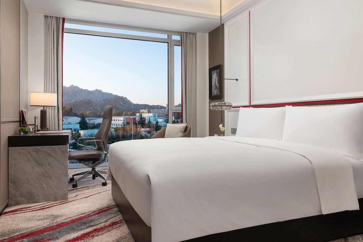 A hotel room with a large bed, a desk, a chair, and a view of mountains and buildings.