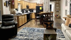 A modern kitchen with wooden cabinets, a dining area with a table and chairs, a black leather chair, and a blue and white rug. - 🛟🎣😎Harbor Lite 2BdrmDreamHouse +Loft! AT the PORT. Peeks of Harbor,River,Ocean! (Brookings)