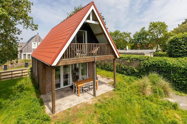 A two-story house with a red roof, a balcony with wooden railings, and a patio area with a table and chairs.