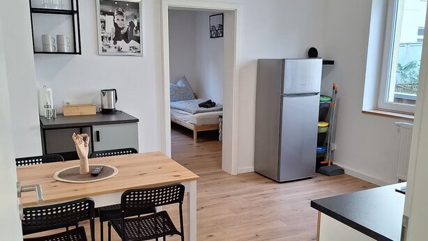 A compact kitchen with a dining area, a bed in the background, and a refrigerator. - Schöne Renovierte, Zentrale 2-zimmer-wohnung in Hagen (Hagen)