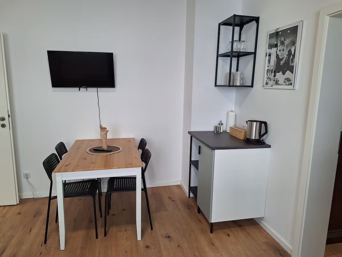 Un espacio habitable compacto con una mesa de comedor de madera, sillas negras, una pequeña kitchenette y un televisor de pantalla plana montado.