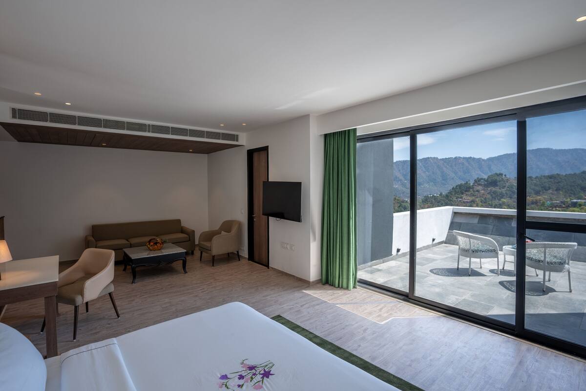 A modern hotel room with a large balcony, a sofa, a dining table, and a TV.