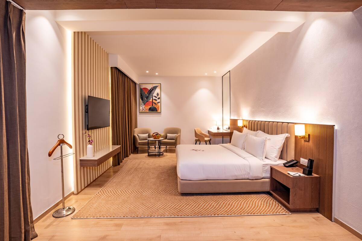 A modern hotel room with a large bed, a desk, a chair, a TV, and a wooden headboard.