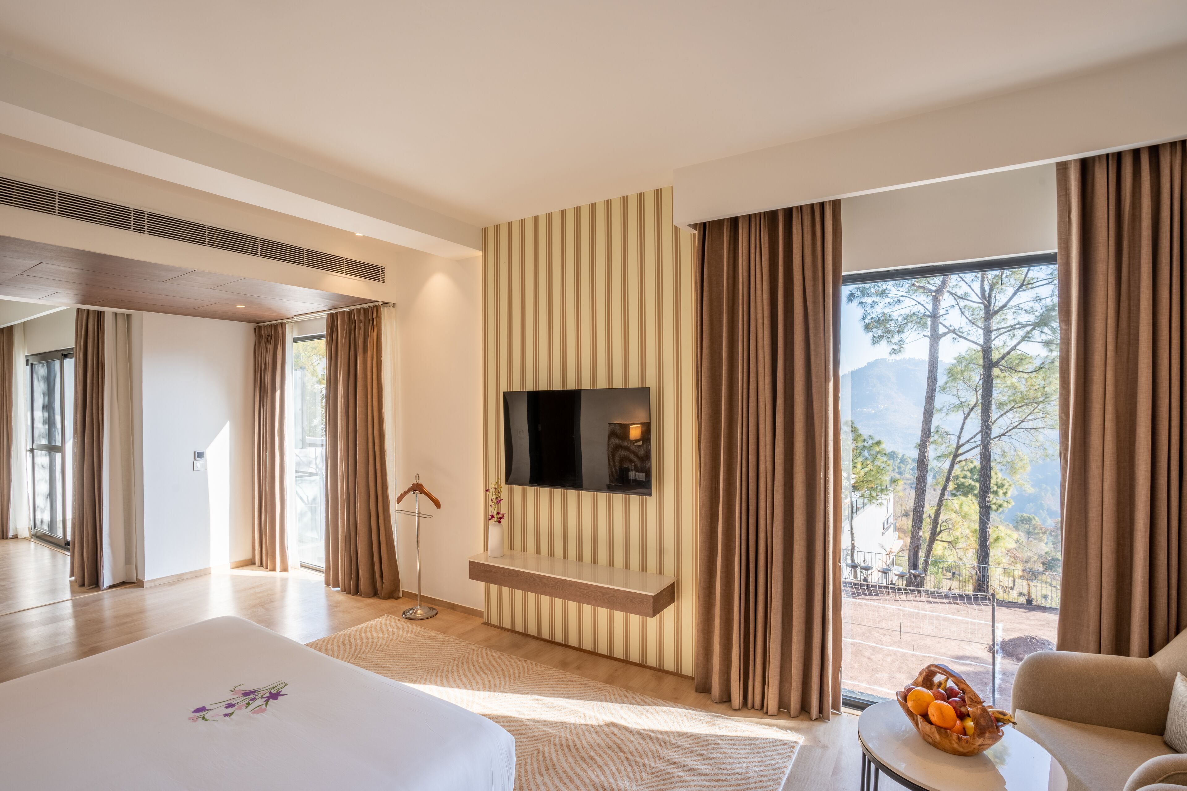 A modern hotel room with a large bed, a TV mounted on the wall, a seating area with a sofa, and a dining table with fruit.