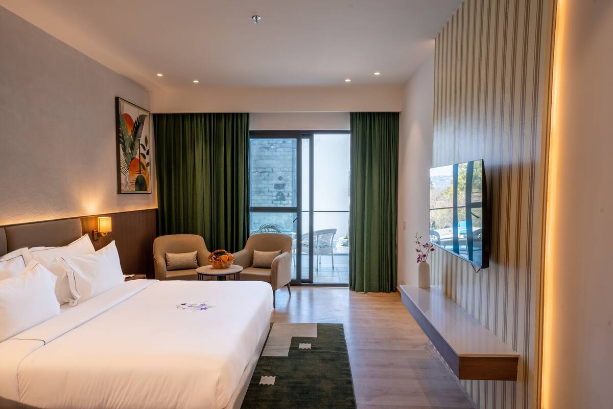 A hotel room with a large bed, a desk, a chair, a TV, and a balcony with curtains.