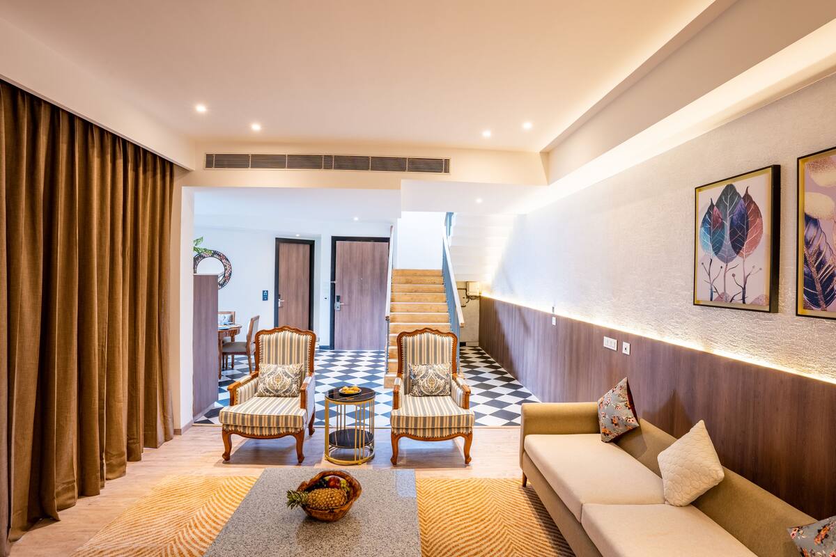 A modern living room with a checkered floor, a sofa, two striped armchairs, a coffee table with fruit, and a staircase in the background.