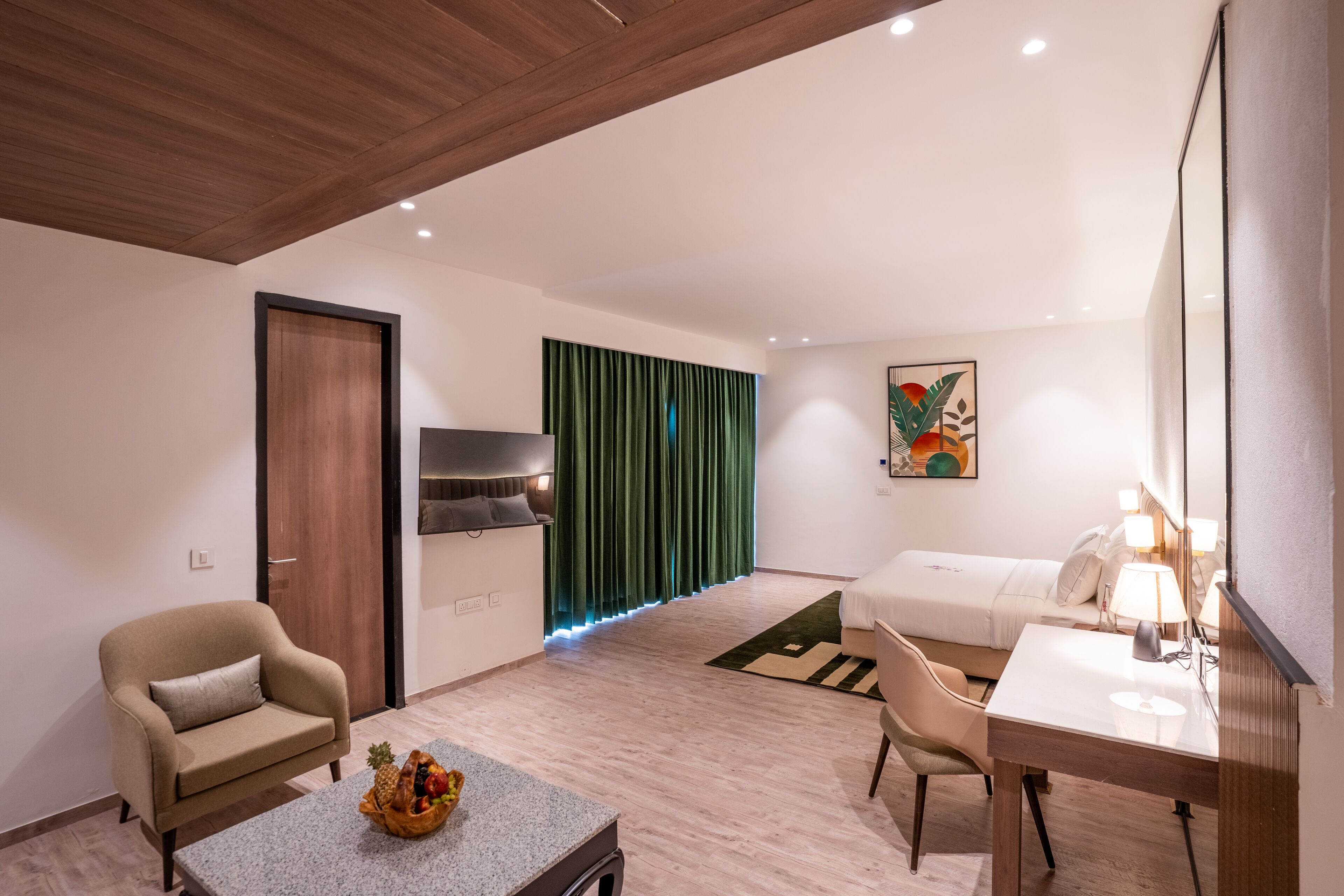 Una habitación de hotel moderna con techo de madera, un espejo grande, un escritorio con lámpara, una cama con cortina verde y una zona de estar con una silla y una mesa con fruta.